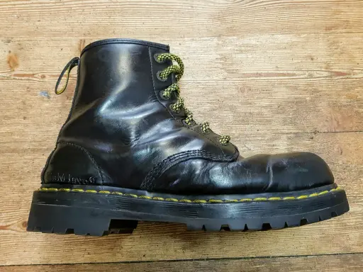 Schwarzer Dr. Martens Stiefel mit gelben Schnürsenkeln und einer neuen, robusten Sohle auf Holzuntergrund.