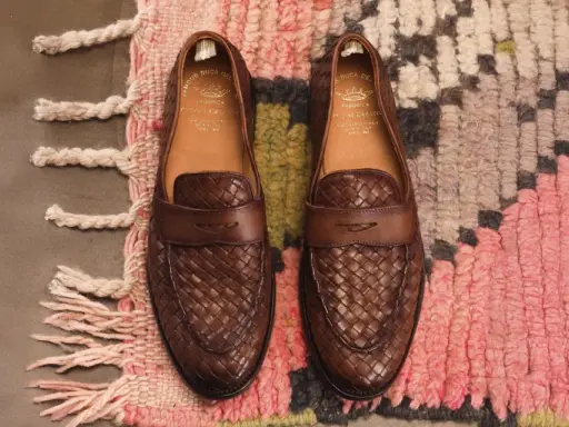 Zwei braune Leder-Loafer mit geflochtenem Muster, Marke Officine Creative, liegen auf einem bunten Teppich.
