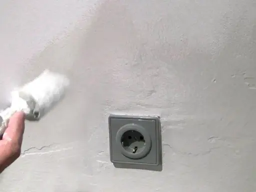 Mit einem Schwamm wird die Wand bearbeitet, um Wasserflecken zu entfernen. Eine Steckdose ist sichtbar.