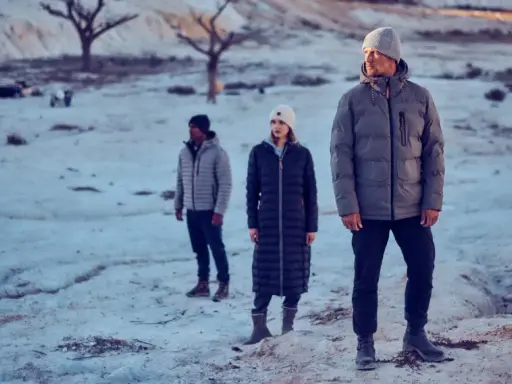 Drei Personen in Winterjacken stehen in einer verschneiten, kargen Landschaft. Die Frage "ab wann Winterjacke" stellt sich bei diesem Anblick von selbst.