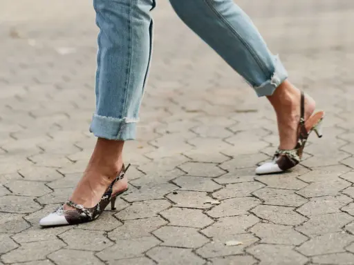 Beine in hochgekrempelten Jeans und Schlangenleder-Pumps auf gepflastertem Untergrund.
