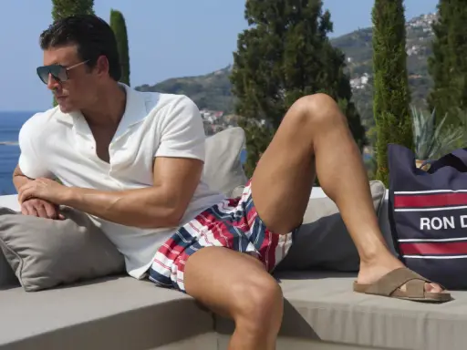 Ein Mann mit Sonnenbrille und gestreifter Badehose entspannt sich auf einer Couch. Neben ihm liegt eine Tasche mit der Aufschrift "RON DORFF".