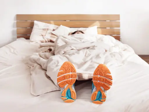 Füße mit Laufschuhen ragen aus einer Decke. Der Wecker klingelt, aber die Person träumt noch vom morgens joggen.