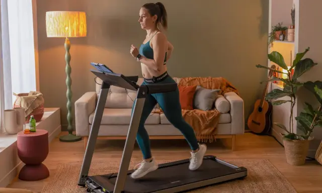 Frau macht Intervalltraining auf Laufband im Wohnzimmer. Sie trägt Sportkleidung und läuft mit Energie.