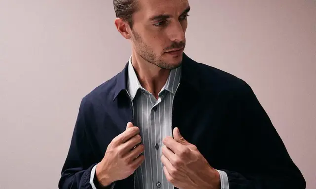 Ein Mann mit Bart und kurz geschnittenem Haar, der eine dunkelblaue Jacke über einem gestreiften Hemd trägt. Er fasst nach dem Reißverschluss, als würde er seine Kleidung für das Hemden waschen vorbereiten.