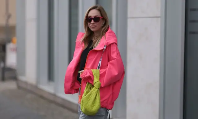Stylische Frau in knallpinker Jacke und Sonnenbrille. Perfekt, um was anziehen bei 15 Grad. Dazu eine grüne Tasche.