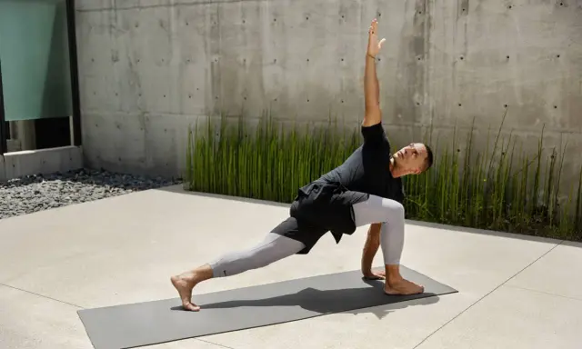 Mann in Yoga-Pose auf Matte vor Betonwand und grünen Pflanzen.