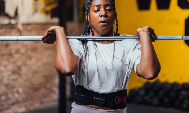 Frau mit geflochtenen Haaren hebt eine Hantel für Jumping Squats. Sie trägt einen Trainingsgürtel.
