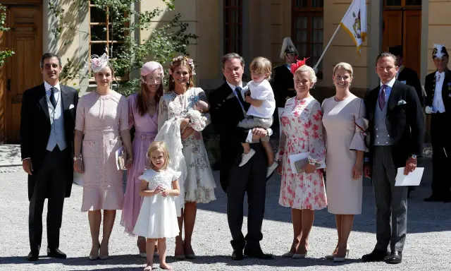 Familie in festlicher Kleidung, die überlegt, was anziehen zur Taufe. Ein Mann trägt ein Baby, Kinder und Frauen in eleganten Kleidern.