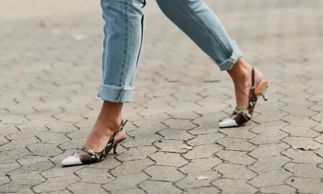 Beine in hochgekrempelten Jeans und Schlangenleder-Pumps auf gepflastertem Untergrund.