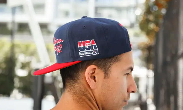 Junger Mann trägt eine dunkelblaue Kappe mit rotem Schirm und "USA Basketball"-Aufdruck, den **cap nach hinten tragen** ist sein Stil.