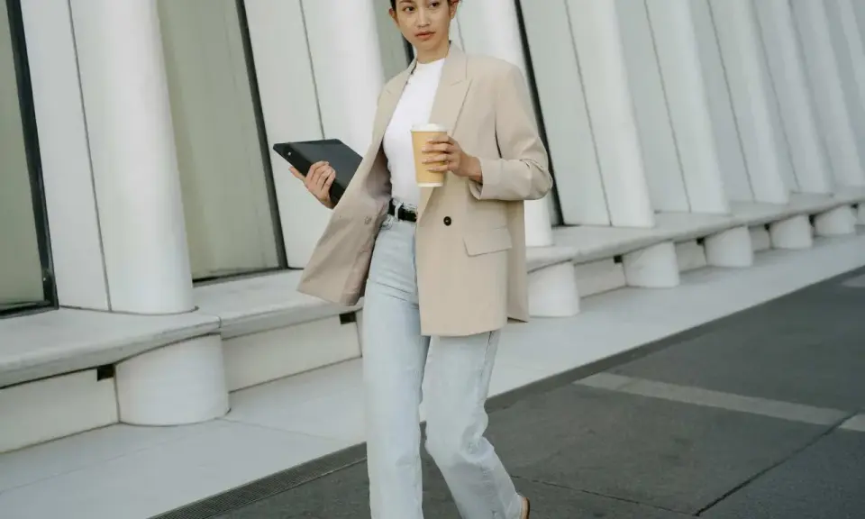 Frau in **smart casual** Kleidung mit Blazer, Jeans und Kaffeebecher geht mit Tablet.