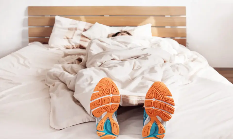 Füße mit Laufschuhen ragen aus einer Decke. Der Wecker klingelt, aber die Person träumt noch vom morgens joggen.