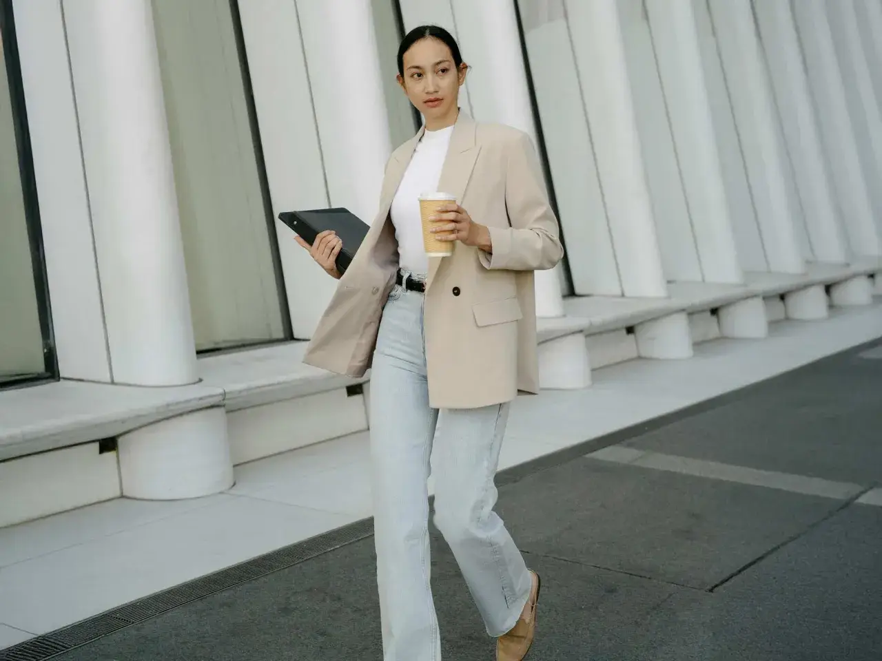Frau in **smart casual** Kleidung mit Blazer, Jeans und Kaffeebecher geht mit Tablet.
