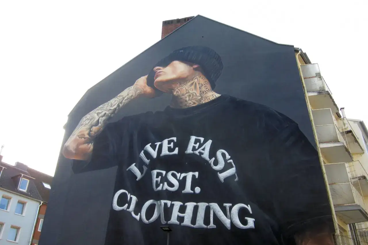 Großes Wandgemälde eines Mannes mit Tattoos, der ein schwarzes T-Shirt mit der Aufschrift