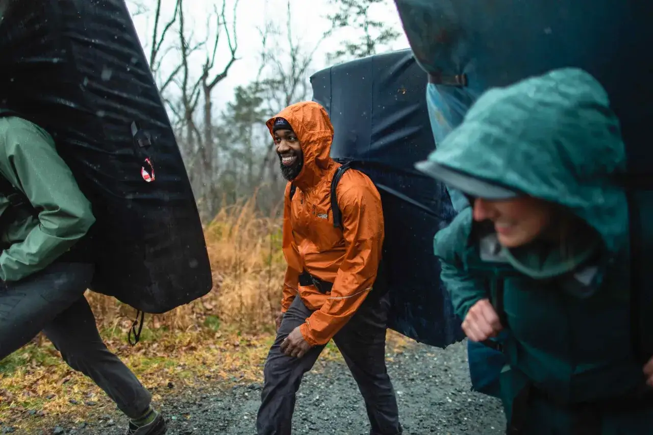 Drei Kletterer tragen dicke Matten durch den Regen. Die orangefarbene Didriksons Jacke des Mannes ist nass, aber er lächelt.