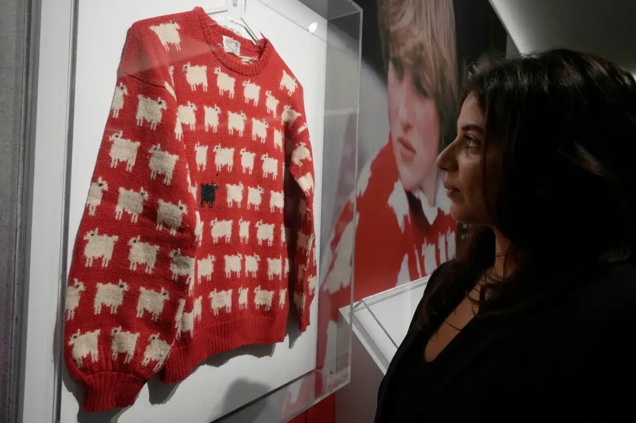 Eine Frau betrachtet den teuersten Pullover der Welt, einen roten Strickpullover mit weißen Schafen und einem schwarzen Schaf.