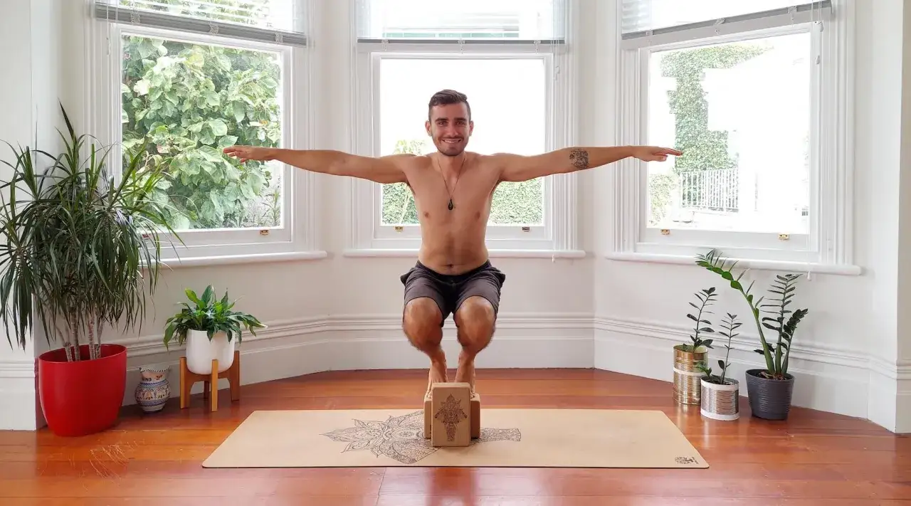 Mann übt Yoga mit Block, steht auf zwei Blöcken in einer Kniebeuge vor einem Fenster.