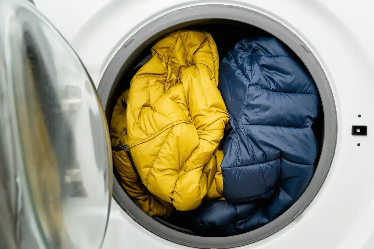 Zwei Daunenjacken, eine gelbe und eine blaue, liegen in der Trommel einer Waschmaschine. Die gelbe Jacke ist auf links gedreht, bereit für die Wäsche.