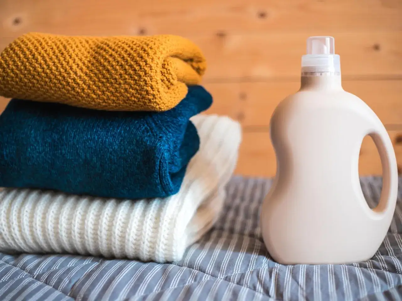 Ein Stapel gestrickter Pullover in Gelb, Blau und Weiß liegt neben einer Flasche Waschmittel. Perfekt für die Wolle waschen.