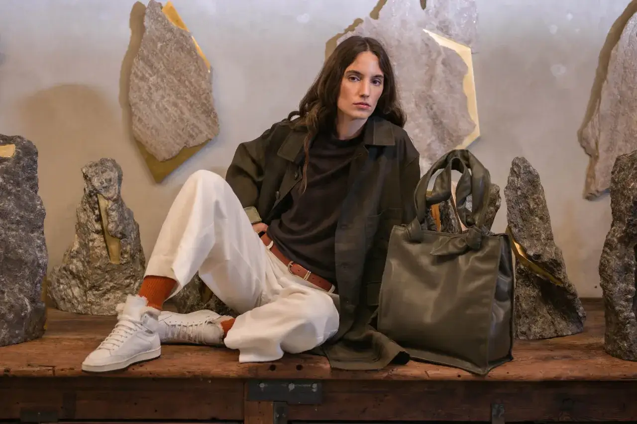 Eine Frau in lässiger Kleidung, inspiriert von Officine Creative, sitzt vor skulpturalen Steinen. Sie trägt eine olivgrüne Jacke, weiße Hose und Sneaker.