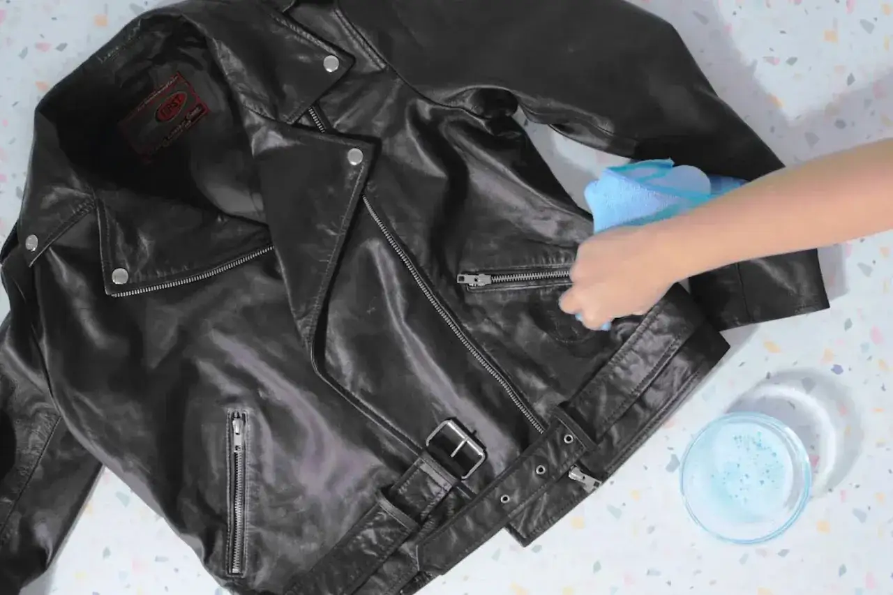 Schwarze Lederjacke reinigen mit einem feuchten Tuch. Eine Hand wischt über die Jacke, daneben eine Schale mit Seifenwasser.
