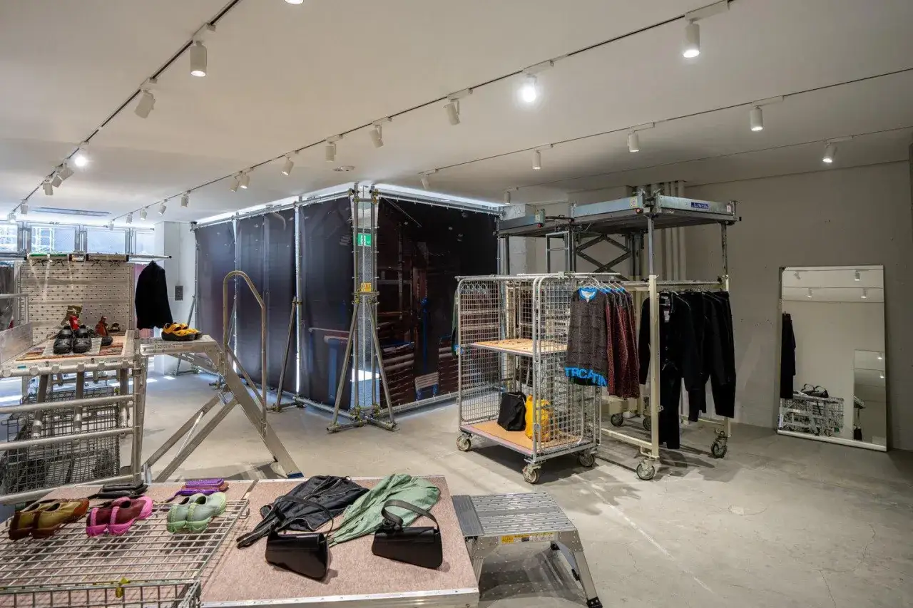Kiko Kostadinov Kollektion in einem minimalistischen Pop-up-Store. Kleidung und Schuhe sind auf Metallständern und Gittern ausgestellt.