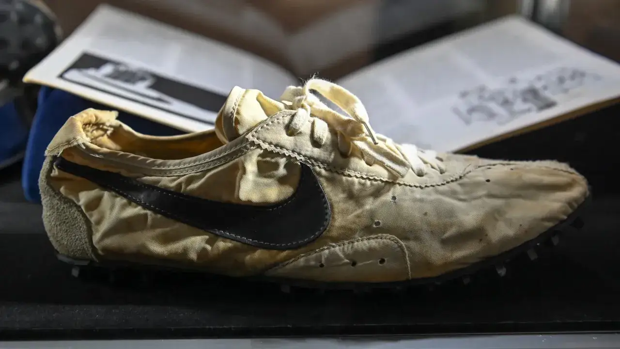 Der erste Nike Schuh von 1972, ein Laufschuh aus hellem Stoff mit schwarzem Swoosh, liegt neben einem geöffneten Buch.