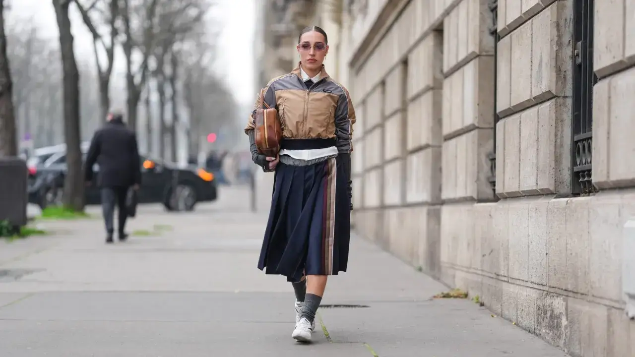 Modische Frau mit dunkelblauem Faltenrock und weißen Sneakern, die perfekt zu ihrem Rock passen. Sie trägt eine braune Jacke und hält eine braune Tasche.