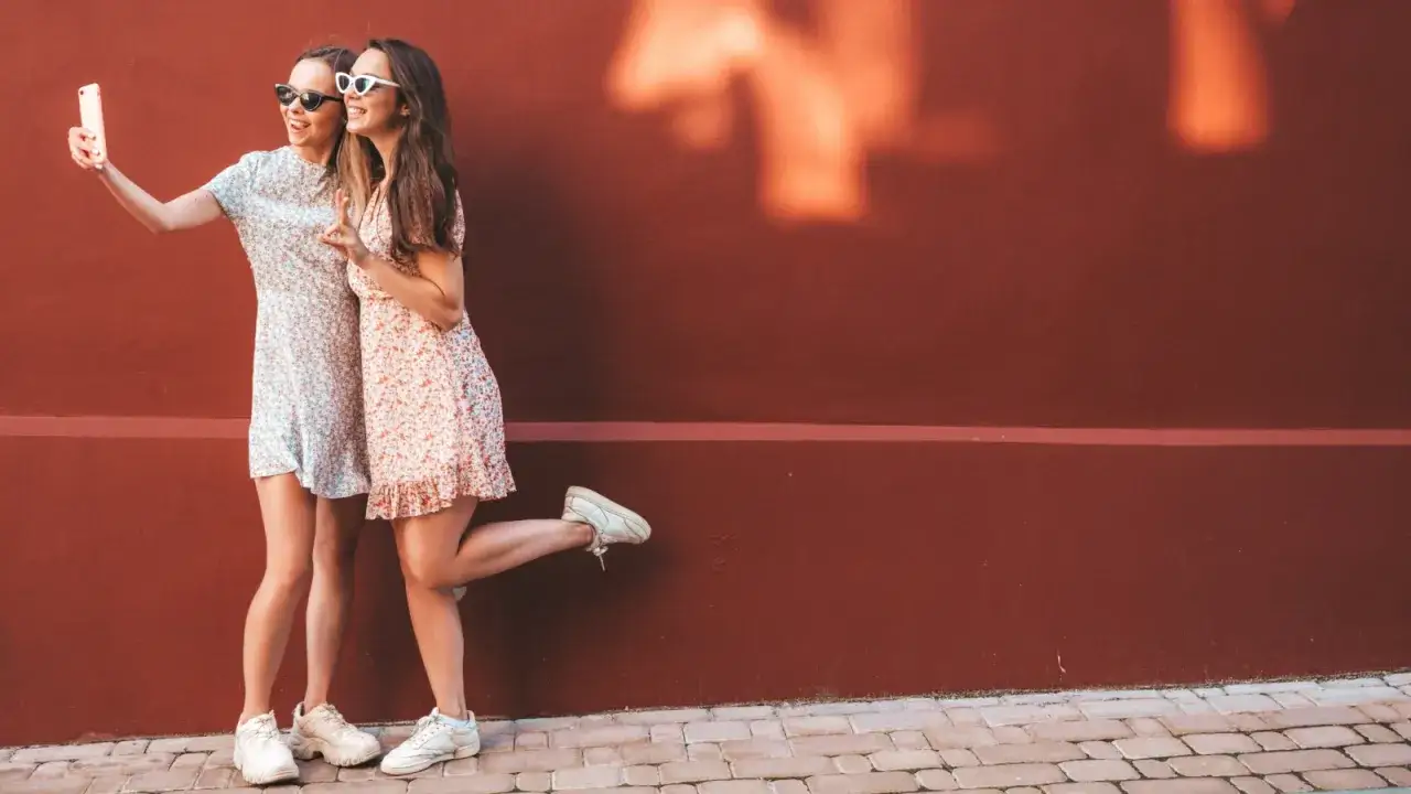 Zwei Freundinnen machen ein Selfie vor einer roten Wand. Beide tragen Sommerkleider und weiße Sneaker zum Kleid.